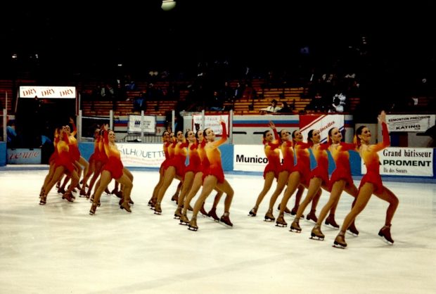 Colour photo showing 16 women in figure skating costumes, performing synchronized movements on an ice rink.