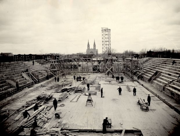 Black-and-white photo of a building construction site, taken from a distance, where about ten men are working on the floor of the building.