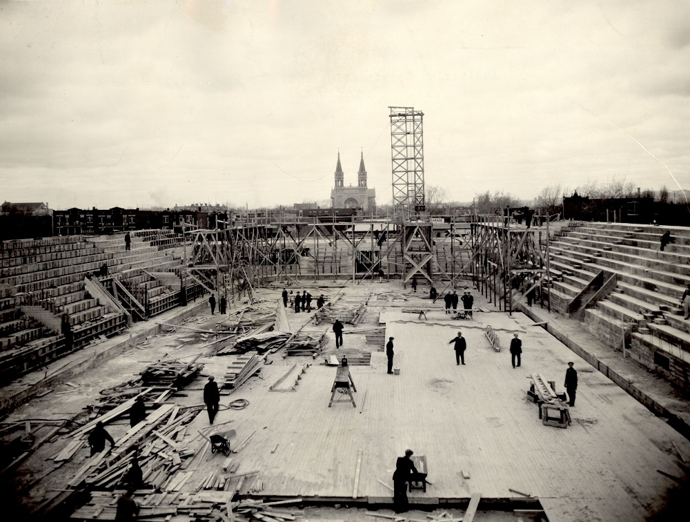 Photographie en noir et blanc d’un chantier de construction d’un bâtiment. La scène est photographiée de loin. Une dizaine d’ouvriers travaillent sur le plancher du bâtiment.