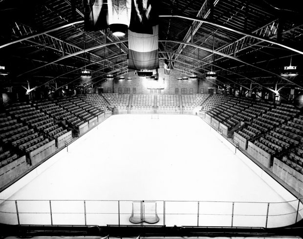 Black-and-white photo of the inside of an arena, where we see a skating rink surrounded by empty stands, as well as hockey nets at each end of the ice surface with flags flying overhead.