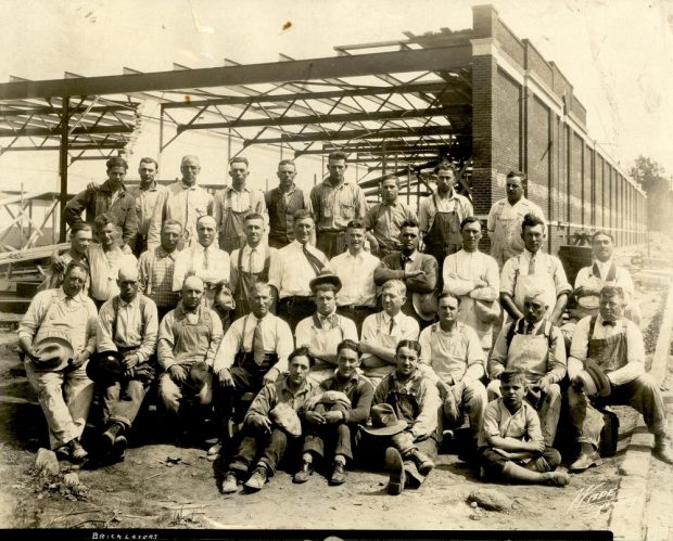 Black-and-white photo showing a group of 33 workers, some seated, others standing, in front of the steel structure of a building under construction. Only the steel structure and a brick wall can be seen.