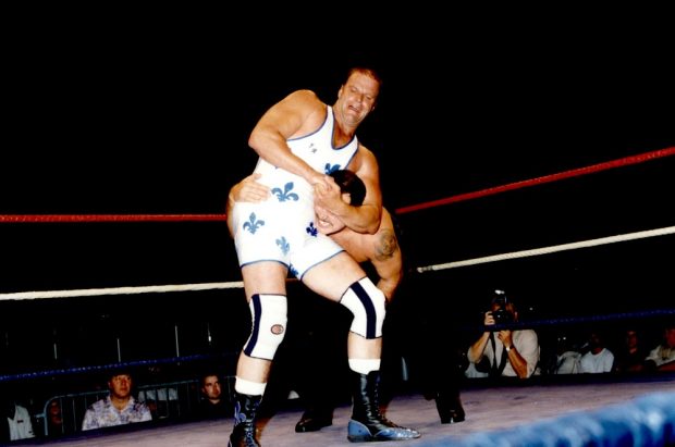 Colour photo showing two men fighting in an arena. The wrestler in the forefront is wearing a white singlet with blue fleurs de lys and kneepads.