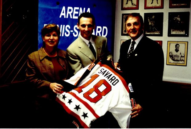 Colour photo of three people: a woman and two men. They hold a hockey sweater with the name Savard, and the number 18 marked on it.