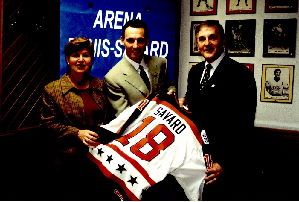 Colour photo of three people: a woman and two men. They hold a hockey sweater with the name "Savard", and the number 18 marked on it.