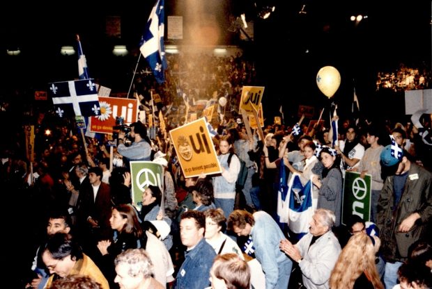 Colour photo showing a crowd of people in the stands holding Québec flags and placards.