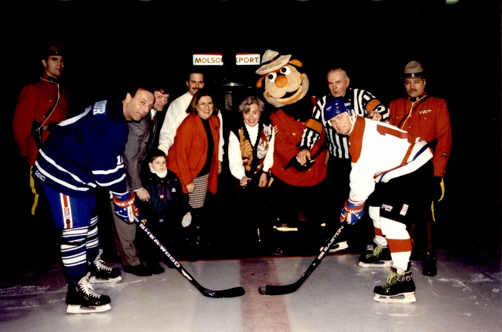 Colour photo showing two hockey players on an ice rink, waiting for the puck to drop. Behind them are five people on a carpet: a child, a man in a referee uniform, two RCMP officers and their mascot.
