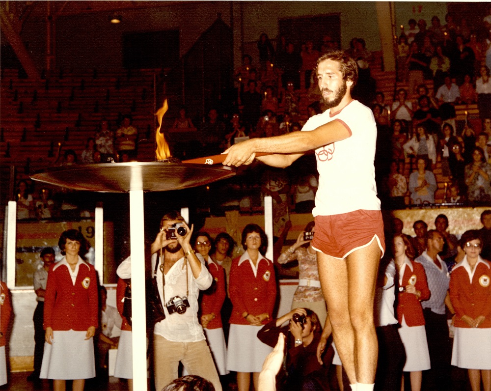 Colour photo of a man wearing orange shorts and a white sweater, holding a lit torch over a receptacle. A crowd can be seen in the background.