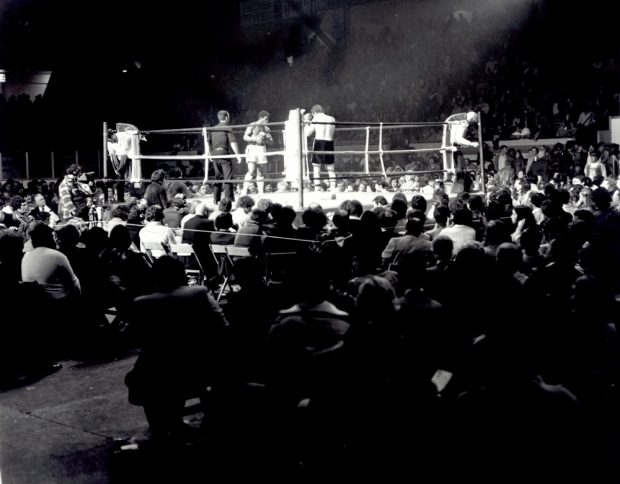 Black-and-white photo showing two men fighting in a boxing arena. A large crowd is cheering on the two athletes.