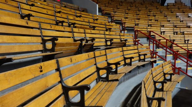 Colour photo showing a close-up of indoor bleachers. The light-brown seating is in the form of two-person benches separated by a black metal armrest. The backs of the seats are numbered 1 to 20.