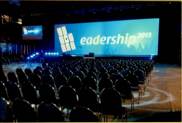 Colour photo showing several rows of empty seats, turned toward a blue screen marked “Leadership 2013”.