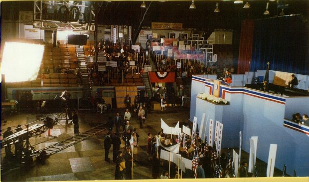 Colour photo showing a big blue podium where one person is standing. Around that podium are people holding signs and movie cameras.