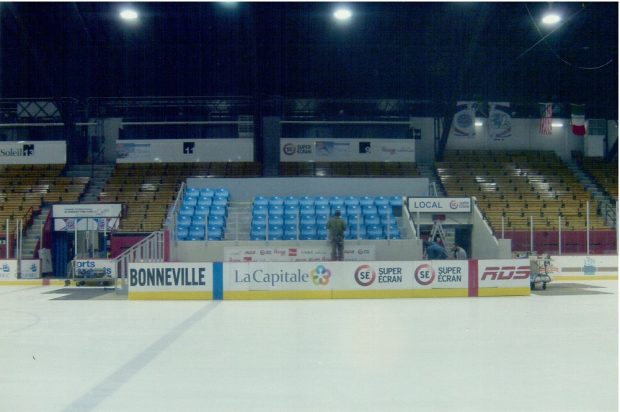 Colour photo showing, in front, a skating rink and its boards, on which several company names and logos are marked. In the middle are a number of rows of blue-coloured seating, surrounded by brown and yellow stands.