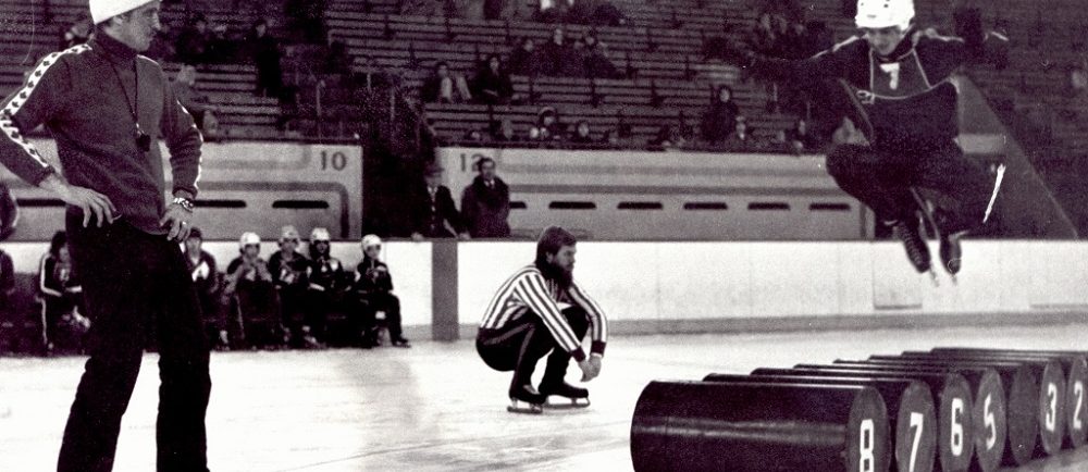 Black-and-white photo of three men on a skating rink. One of them is jumping over some barrels.