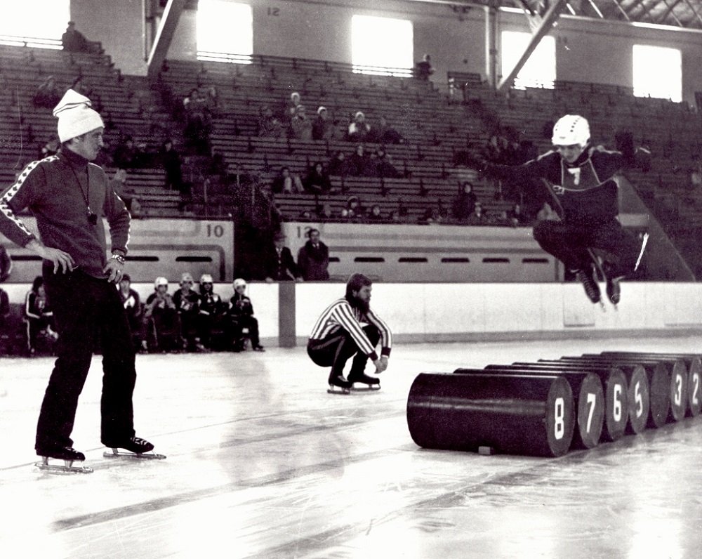 Black-and-white photo of three men on a skating rink. One of them is jumping over some barrels.