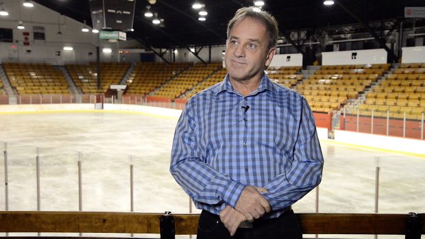 Colour photo of a man standing up. Behind him, there are a skatink rink and stands.