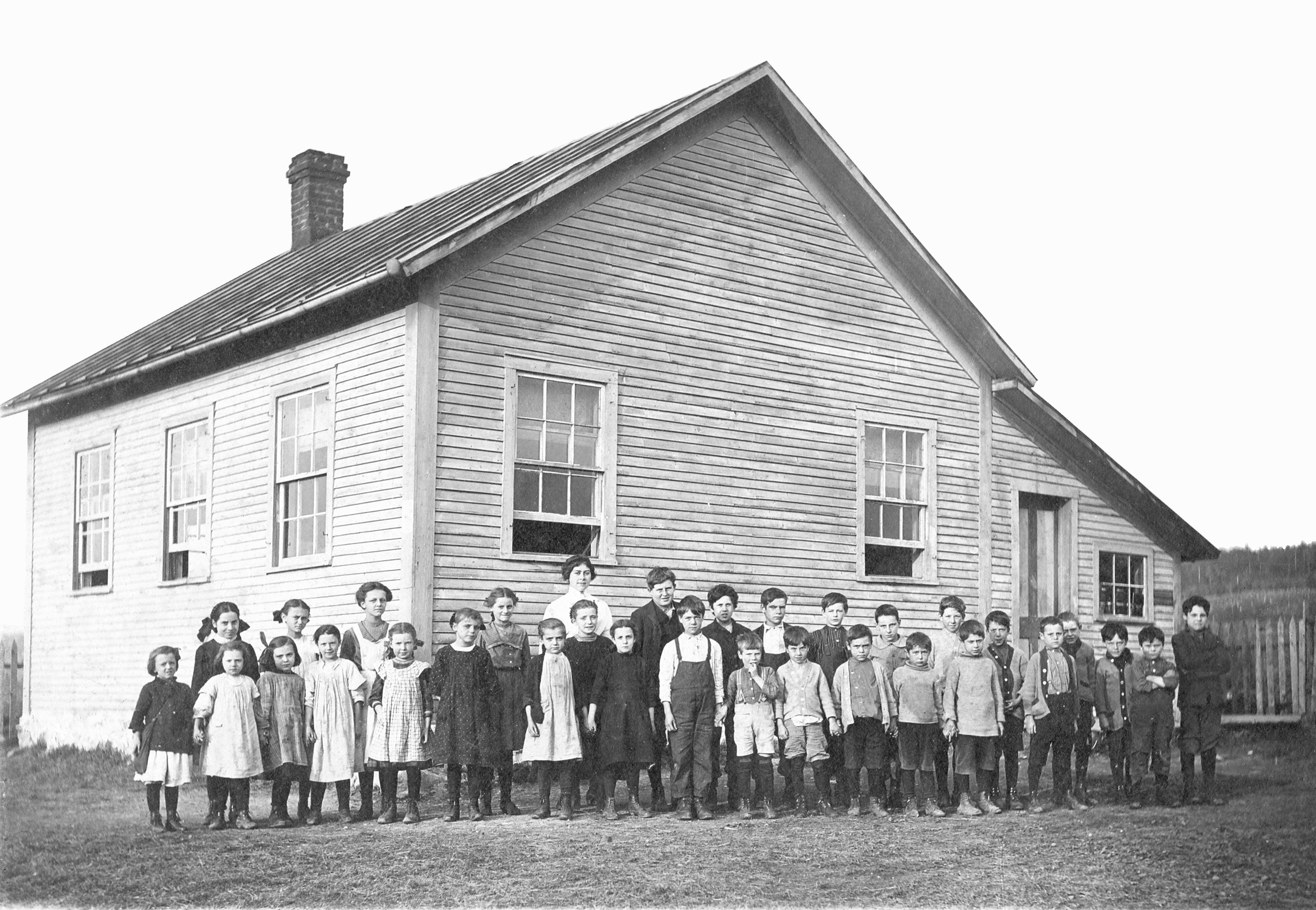 Twenty-nine school children of all ages pose with their teacher in front of a small white woodenbuilding.