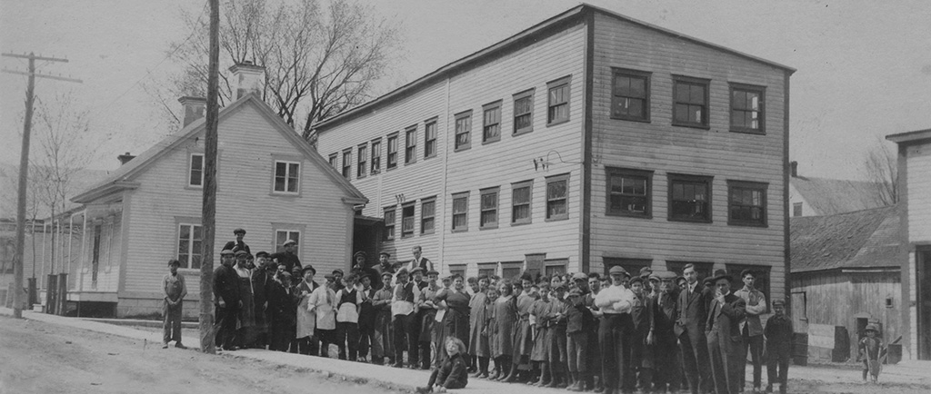 The picture shows employees assembled in front of the Charron factory. It is a three-storey wooden structure, built behind a house on St-Antoine Street.