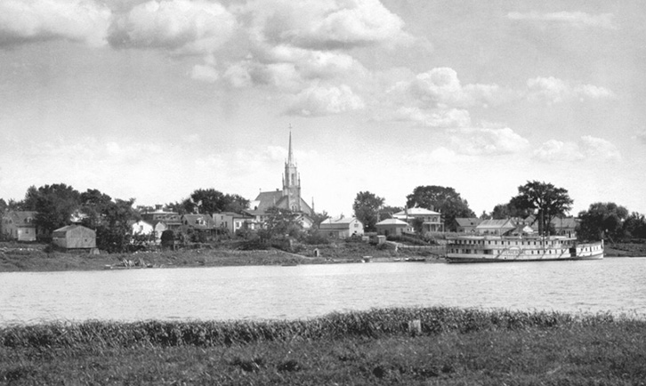 The photo shows a view of the town of Contrecœur from the island that faces the church. We can see the Terrebonne steamboat berthed at the dock.