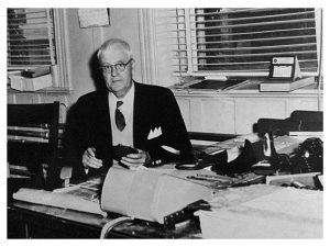 La photo montre  Joseph Papin III dans son bureau.  Il semble inspecter une paire de chaussures.