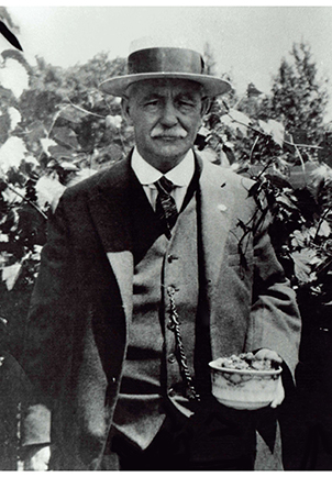 Photo of Joseph Papin II dressed in a suit in a courtyard. He wears a hat and appears to be holding bowl of fruit in his left hand.