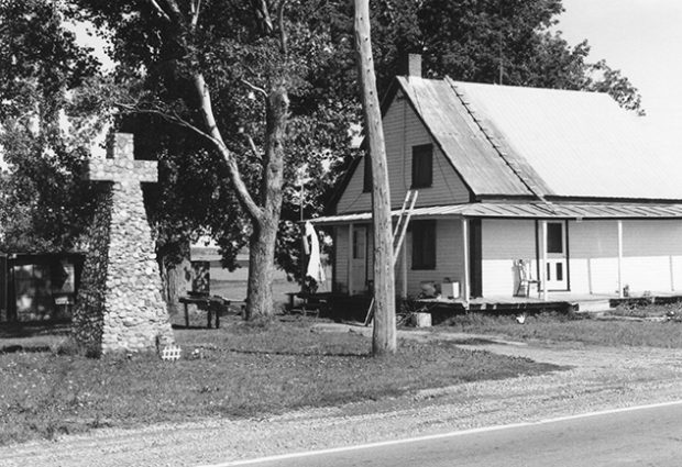 This photo shows a small, white wooden houses located on the edge of the St. Lawrence River. Next to it, a stone cross was erected to indicate the location of the first original Contrecœur church.