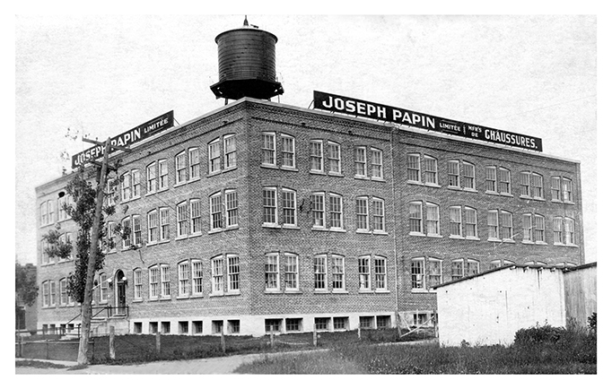 La photo montre la manufacture Joseph Papin ltée, après l’agrandissement de 1924. C’est un imposant bâtiment en brique de quatre étages sur lequel trônent un château d’eau et deux immenses enseignes.  