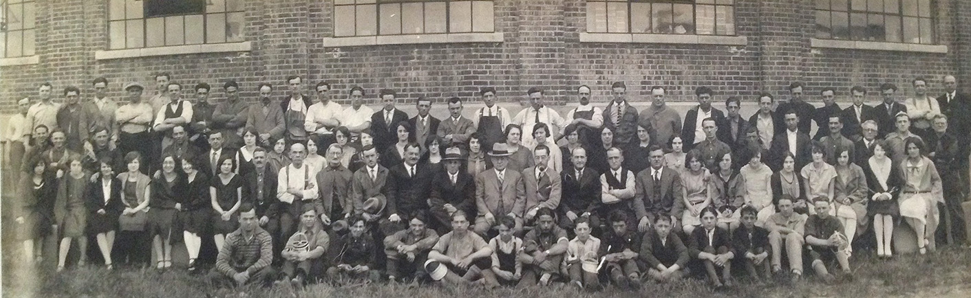 The picture, taken in front of the factory, shows Albert Charron factory employees in 1928. They number close to a hundred: men, women and children. We see the brick wall and large windows in the background.