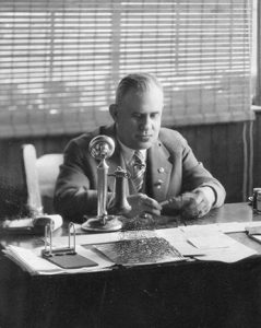 Photo montrant Albert Charron assis à son bureau devant une grande fenêtre.  Il est vêtu d’un complet-veston très chic.  Sur son bureau, on retrouve un téléphone métallique vertical.