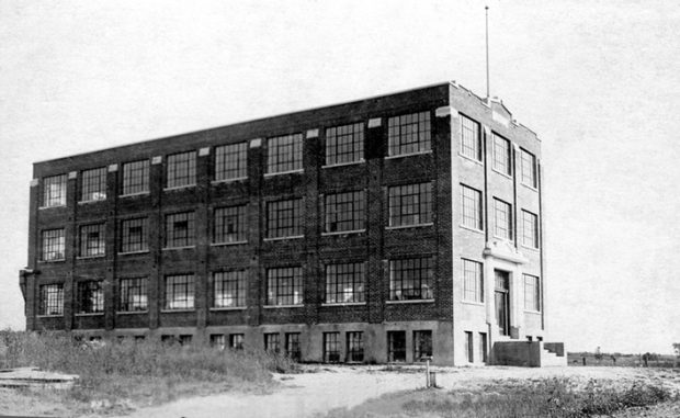 This picture shows the Charron Factory shortly after its construction in 1924. A dirt road leads to the main entrance. No other structure can be seen nearby. It is a brick building with four storeys that appears to be located in the middle of a field.