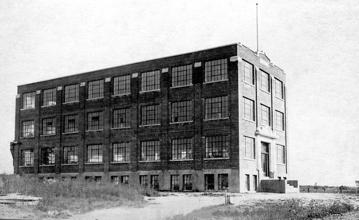 This picture shows the Charron Factory shortly after its construction in 1924. A dirt road leads to the main entrance. No other structure can be seen nearby. It is a brick building with four storeys that appears to be located in the middle of a field.