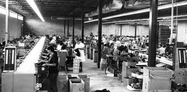 The picture shows the sewing department. We see approximately forty workers spread out on either side of two long conveyors that cut across the room.