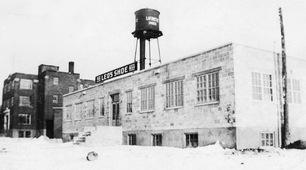 Picture showing the Leo’s shoe Reg’d plant. It is a two-storey, grey brick building. Behind it, we see the Lafayette plant with its water tower.