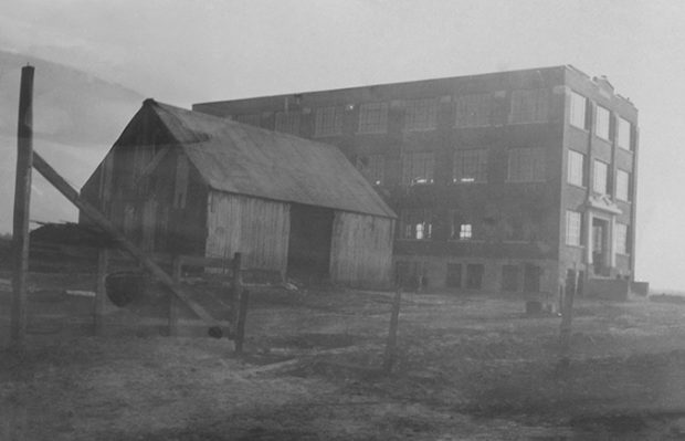 The picture shows the Charron Factory, shortly after it was built in 1924. It is a large, four-storey brick building. No street services the area yet. We see only a wooden barn next to the plant.