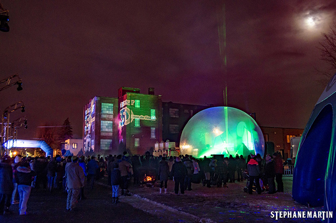 This picture shows the Lafayette factory lit up by projectors on the 350th anniversary of the city of Contrecœur. We see a futuristic dome in front of the old factory and hundreds of people assembled.