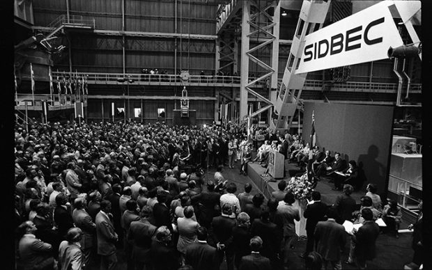 The picture was taken inside the Sidbec factory during its inauguration. On a stage set up for that purpose, we see several dignitaries, including Prime Minister Robert Bourassa at the mike. On the floor, hundreds of people have gathered to participate in the event.