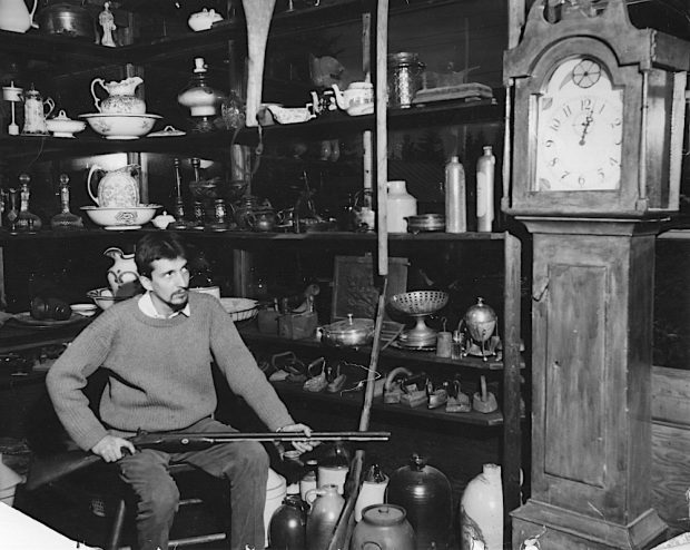 Black and white photo of the Butte antique store. Gilles Mathieu is seated and holds an old rifle. A grandfather clock predominates in front of shelves on which are presented antiques.