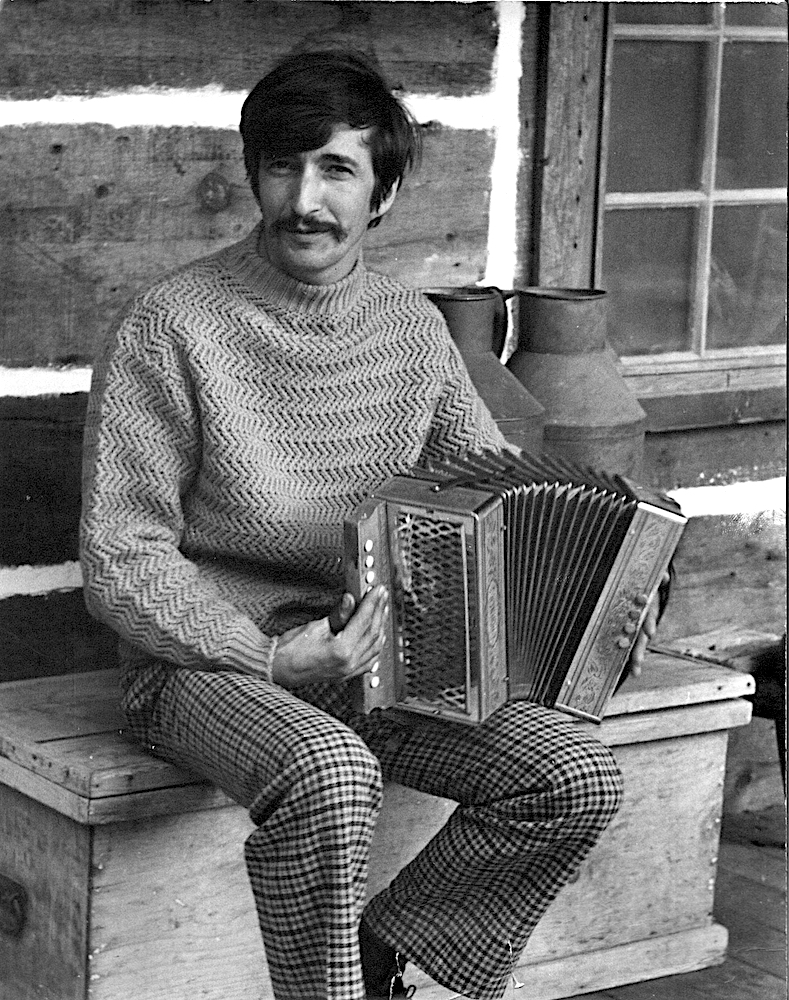 Black and white photo of Gilles Mathieu sitting on a wooden bench on the balcony of his house on the site of La Butte and playing the accordion.