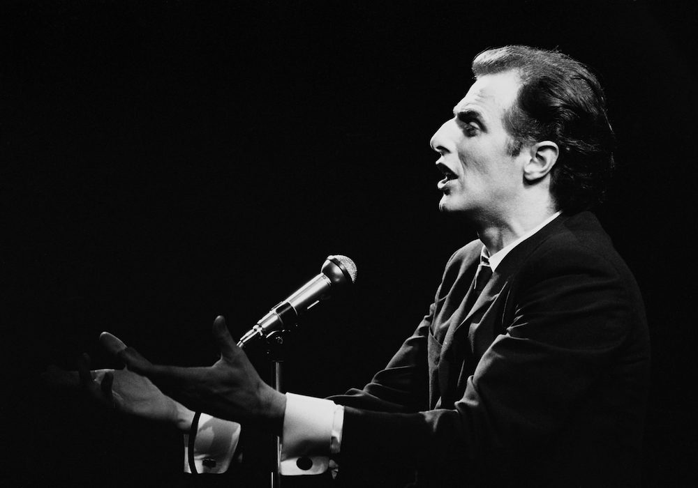 Black and white photo of Gilles Vigneault with his mouth open and his hands outstretched in front of his microphone.