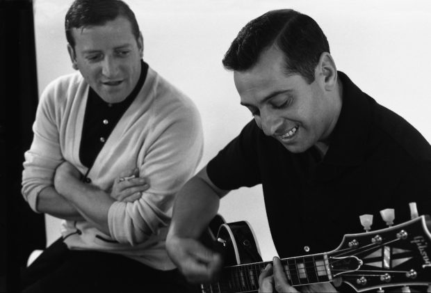 Black and white photo of the Jérolas. Jérôme Lemay on guitar is listened to by Jean Lapointe.