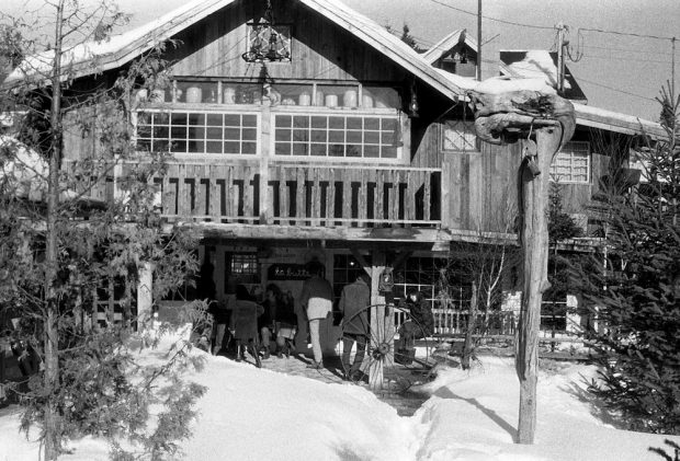 Black and white photo of La Butte's main entrance in winter outside. On its old barn wood walls, a large gallery runs alongside paneled windows.