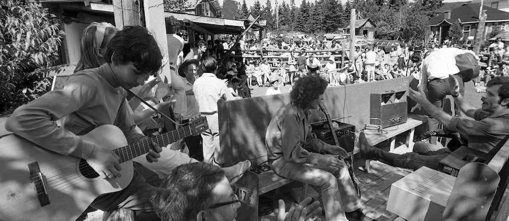 Black and white photo of about 100 spectators attending an outdoor performance at La Butte. At the top of the picture, the feet of people sitting on part of the roof of La Butte can be seen.