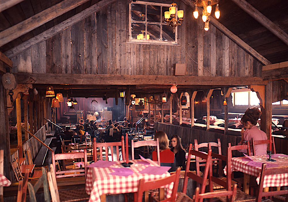 Colour photo of the main room of La Butte. Several tables with checkered tablecloths are set up in front of the main stage.