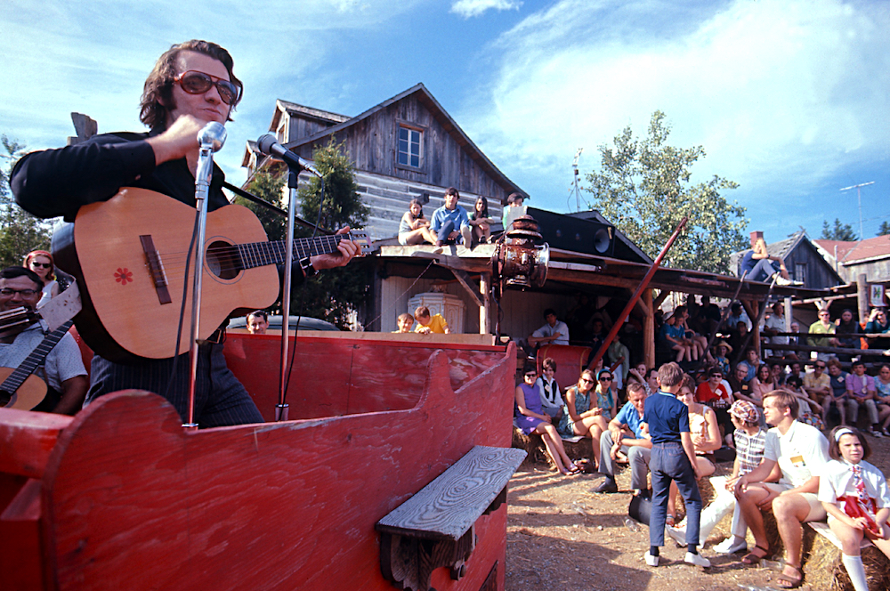 Color photo of Jean-Guy Moreau on guitar, sunglasses on his nose performing outside at La Butte in front of a crowd.