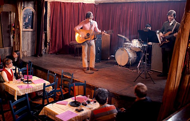 Colour photo of two young musicians on the stage of La Butte, one standing with a guitar, the other sitting behind the drums. Spectators sitting at tables in the room are listening to them and watching.