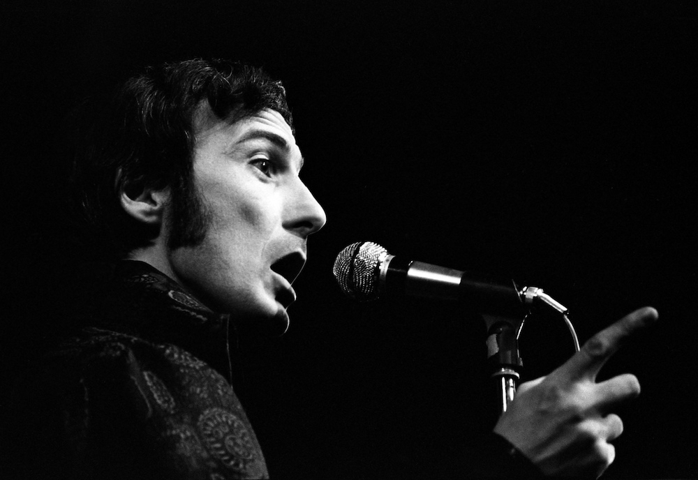 Black and white photo of Yvon Deschamps performing with his mouth open and pointing at the audience in front of his microphone.