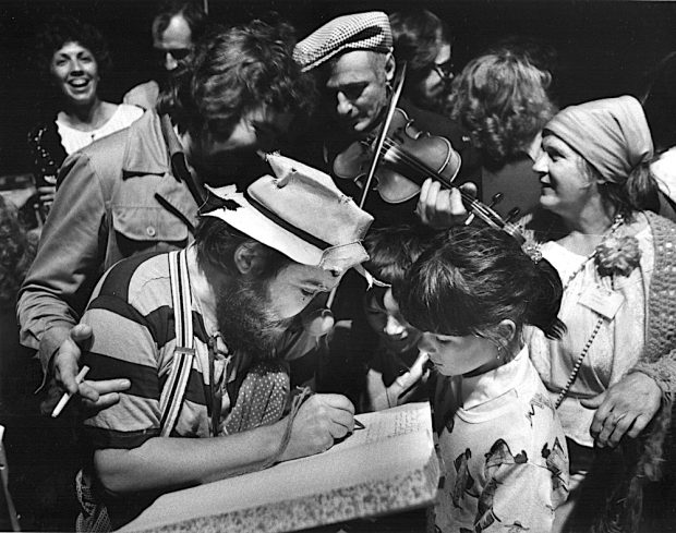 Black and white photo of buskers at La Butte. A group of children surrounds a clown, a fiddler and Gilles Mathieu.