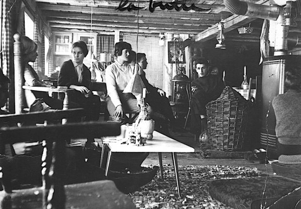 Black and white photo of half a dozen customers sitting in the first Butte building. A candle stuck in the neck of a Chianti bottle sits on a coffee table. The beams are exposed.