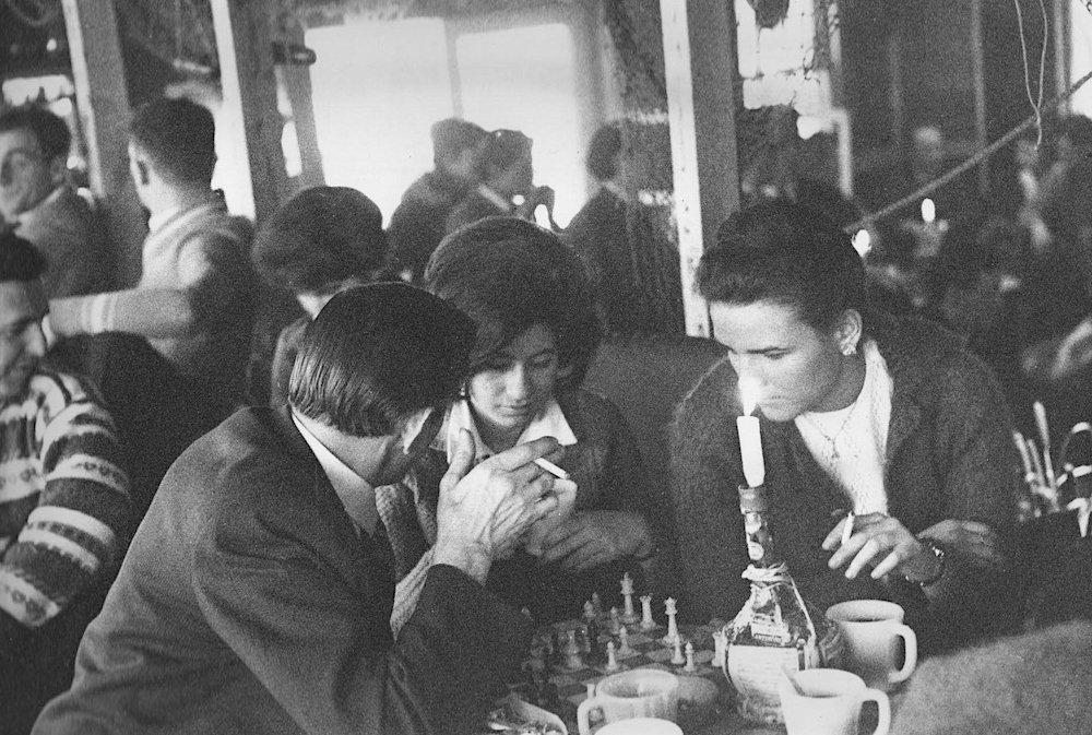 Black and white photo of two men and a woman playing chess on a table at La Butte surrounded by several other people.