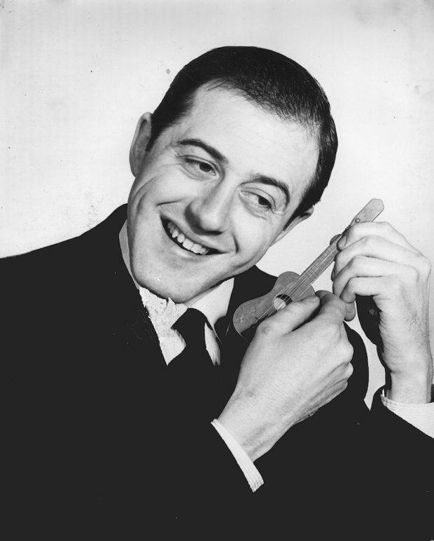 Black and white photo of Raymond Lévesque in a jacket and tie listening to the sound of a miniature guitar he is holding in his hands.