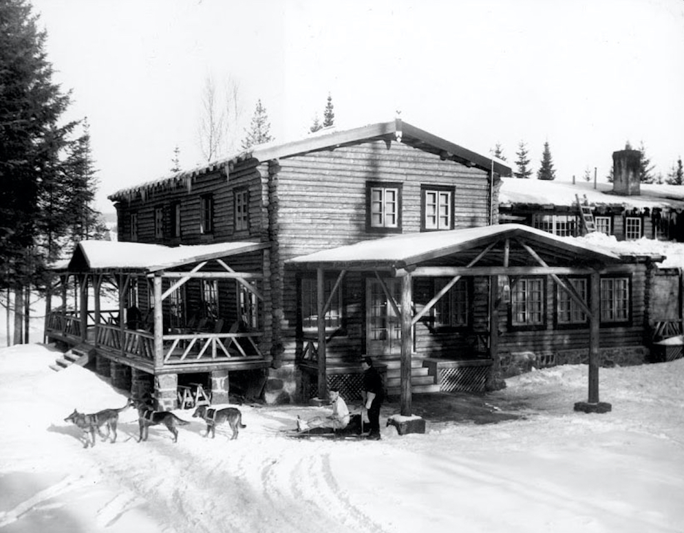 Black and white photo of Hôtel La Sapinière in the snow in the winter of 1936-1937. In front of the log building is a sled with two people and three sled dogs.
